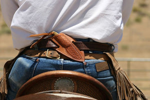 Close-up of a person wearing a white shirt, blue jeans, and a brown leather chaps belt with fringe, standing outdoors near a horse saddle.