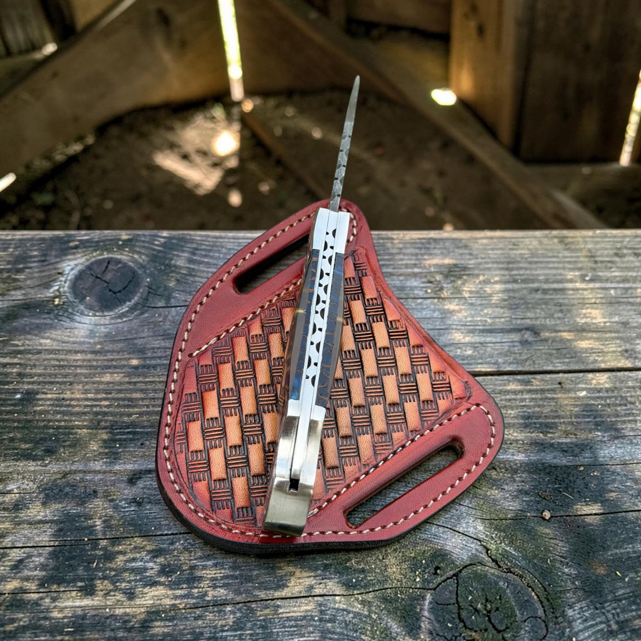 A folding pocket knife with a silver blade and handle, resting on a reddish-brown woven leather sheath over a weathered wooden surface.