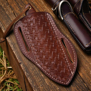 A dark brown embossed leather sheath with a woven basket-like pattern and contrast stitching, shown on a rustic wooden surface next to another leather sheath.