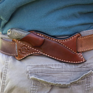 A close-up of a brown leather knife sheath worn over a belt on a person's waist, with part of a blue shirt and light gray jeans visible.