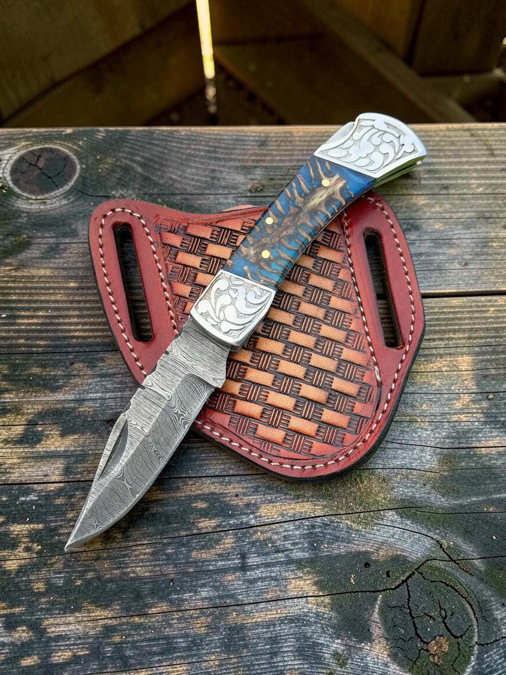A fixed-blade knife with a patterned blue and gold handle, silver-toned engraved metal blade and bolster, resting on a brown woven sheath over a weathered wooden surface.