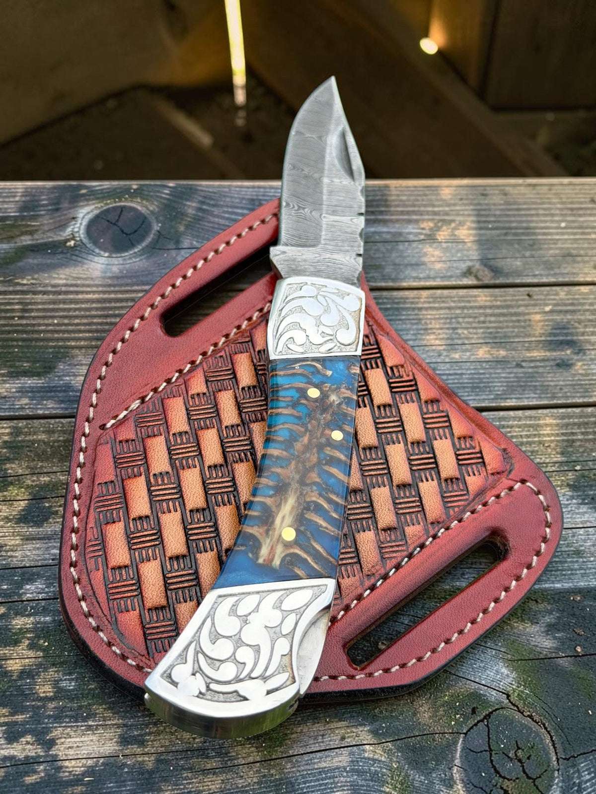 A custom fixed-blade knife with a textured silver blade, ornate silver-toned metal guard and bolster, and a blue marbled handle with gold flecks, resting on its reddish-brown woven leather sheath over a rustic wooden surface.