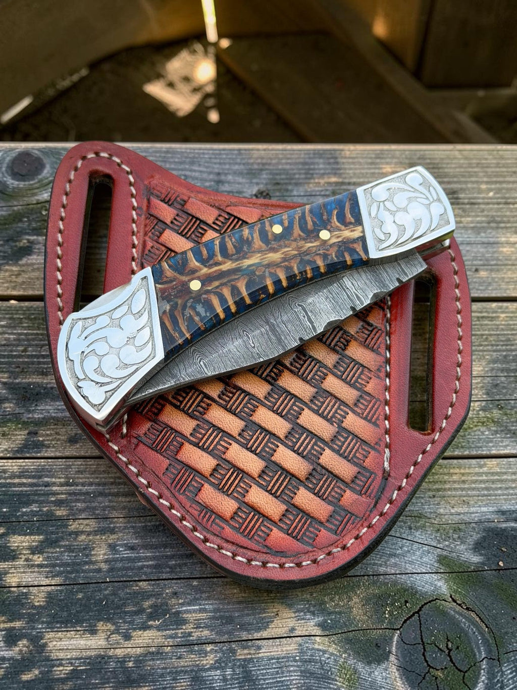 A folding pocket knife with a patterned wood handle and silver-toned metal end pieces, resting on a reddish-brown leather sheath over a weathered wooden surface.