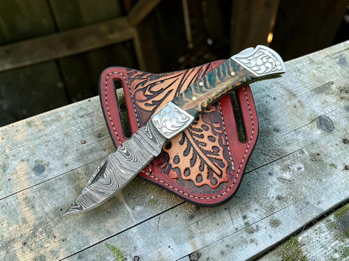 A custom knife with a patterned steel blade and carved handle, resting on a red leather sheath with floral tooling, placed on a weathered wooden surface.
