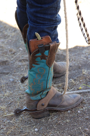 A close-up of a person's lower legs wearing turquoise and brown western cowboy boots with dark blue jeans, standing outdoors on dirt ground next to a rope.