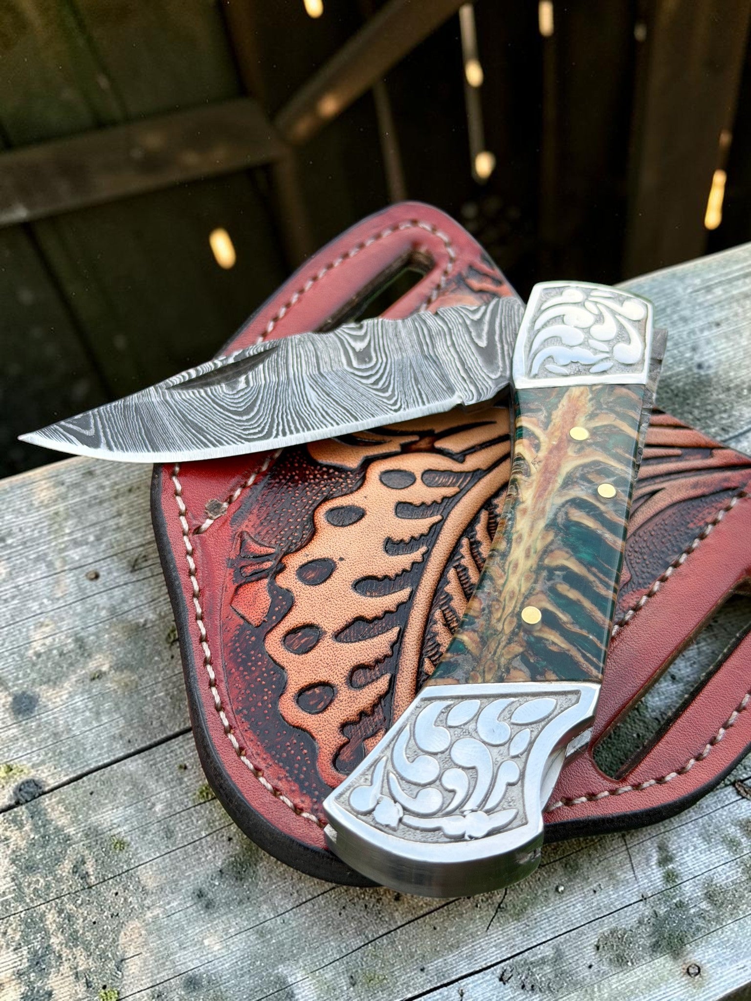 A custom handmade folding knife with a Damascus steel blade and a carved handle, resting on a red leather sheath on a weathered wooden surface.
