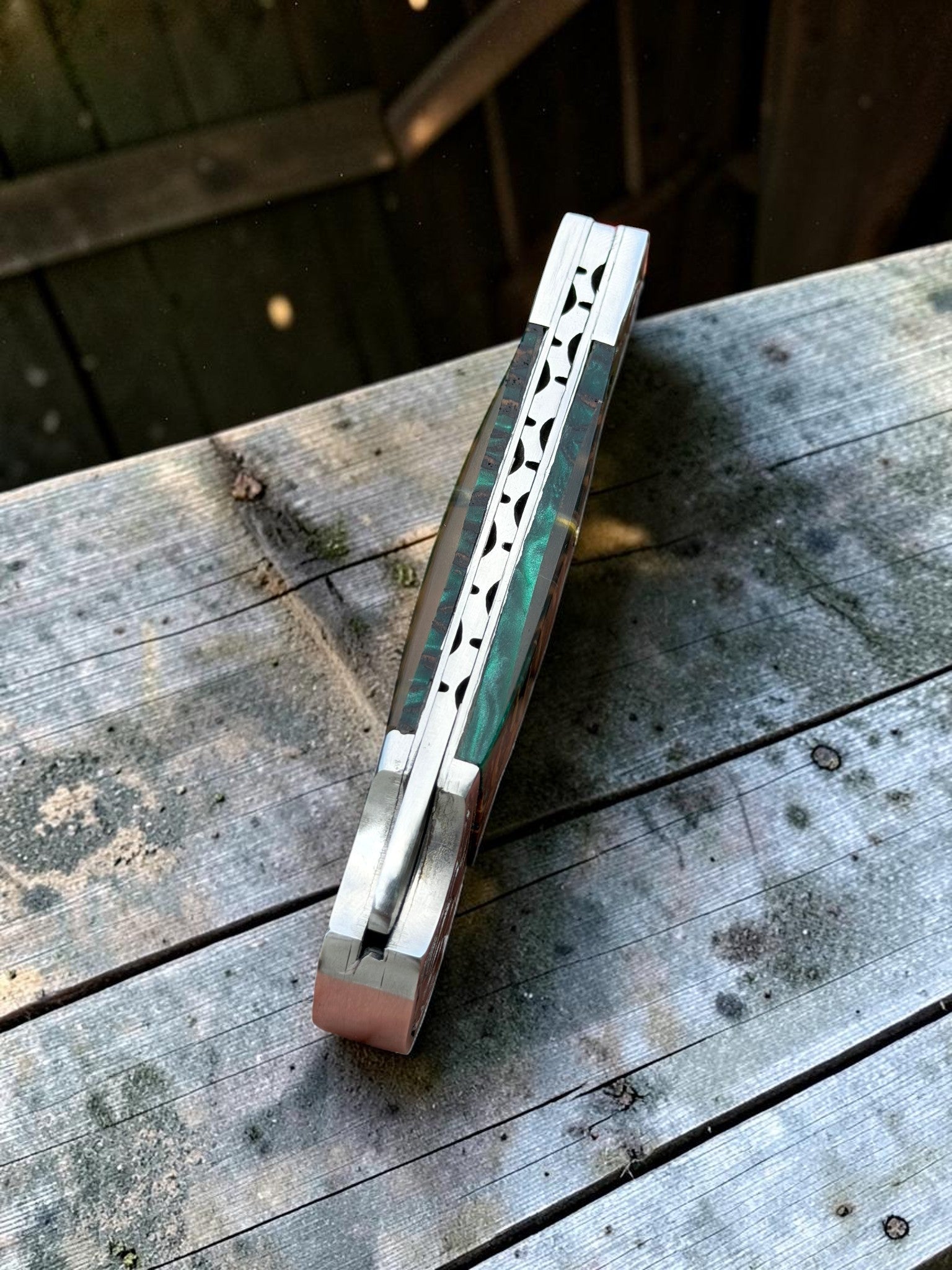 A folding pocket knife with a silver-toned metal frame and a green inlay handle, shown open on a weathered wooden deck.
