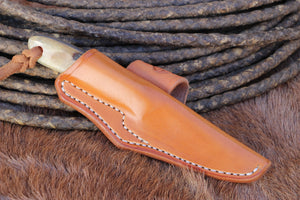 A tan leather knife sheath holding a knife with a light-colored handle, styled on brown fur with a coiled dark rope in the background.