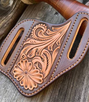 A close-up of a brown tooled leather axe sheath with floral and scroll embossed patterns, resting on a rustic wooden surface.